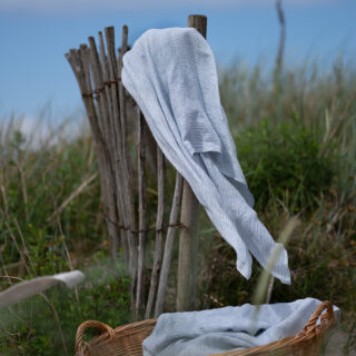 Linen towel from Vävaren i Båstad draped over a wooden fence by the beach, with a woven basket and straw hat in the foreground.